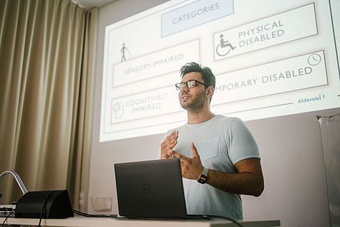 Deniz hält einen Vortrag. Vor ihm ein Laptop und hinter ihm seine Präsentation.