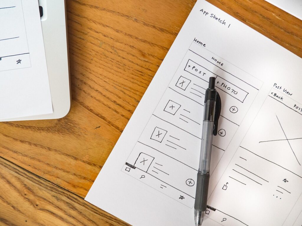Papier mit Skizzen einer App-Oberfläche, darunter ein Stift auf einem Holztisch.